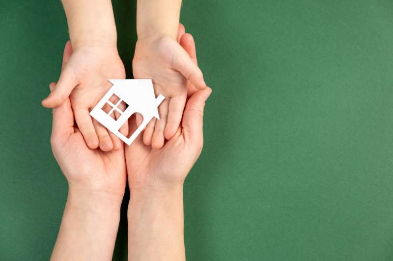 Close up of female and child hands holding white paper house on green background. Family home and real estate concept. Flat lay, copy space